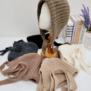 May include: A collection of knitted bonnets in various colors, including brown, gray, and beige, with long ties. One bonnet is displayed on a mannequin head. The bonnets are arranged on a white surface, with books and a small paper bag in the background.