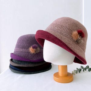 May include: A stack of four felt hats with a contrasting colored band around the bottom. The top hat is a light brown with a burgundy band and a small brown and black pom-pom. The second hat is purple with a purple band and a small brown and black pom-pom.
