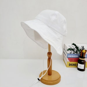 May include: A white bucket hat with a white drawstring. The hat is displayed on a wooden stand.