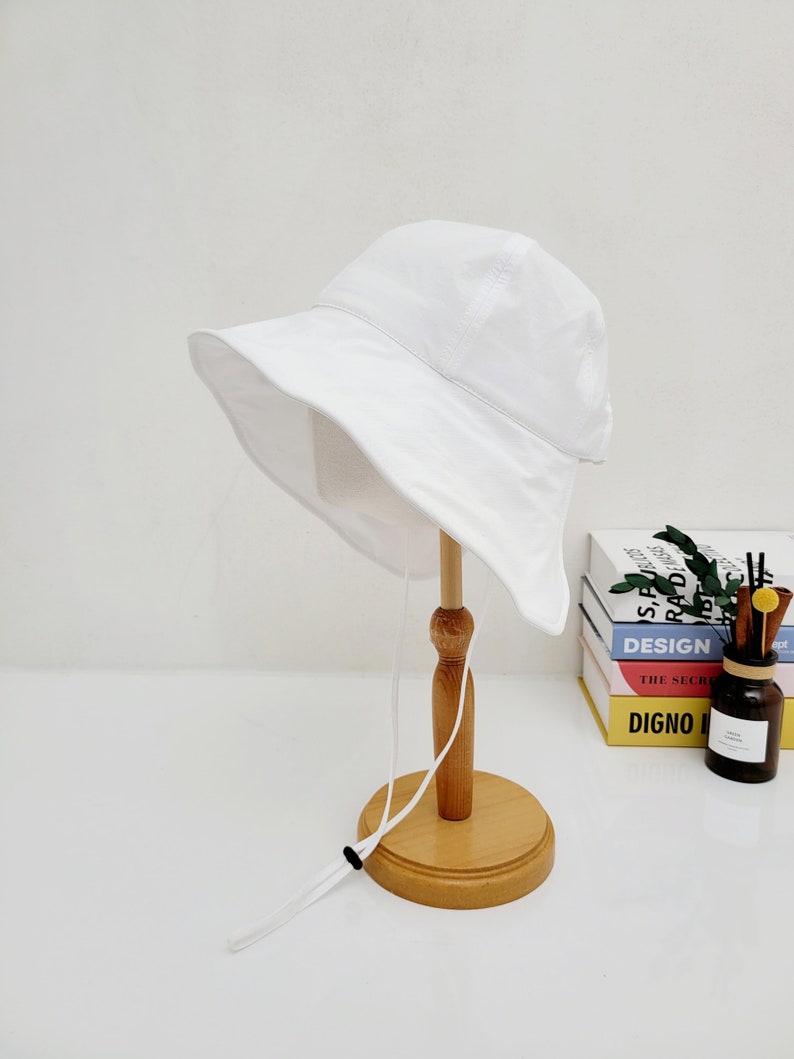 May include: A white bucket hat with a white drawstring. The hat is displayed on a wooden stand.