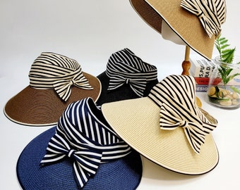 May include: Five wide-brimmed sun hats in various colors: brown, black, navy blue, and beige. Each hat features a striped bow and a contrasting trim. The hats are made of woven material, perfect for sun protection.