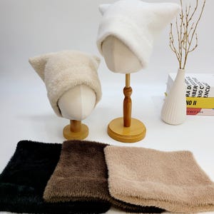 May include: Two cat ear hats, one cream and one white, displayed on mannequin heads. Below are three matching neck warmers in black, brown, and tan. The hats and neck warmers are made of a soft, fuzzy material.