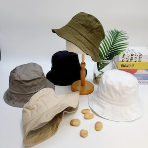 May include: Four bucket hats in different colors: olive green, black, gray, and beige. The hats are arranged on a wooden stand and a white surface.