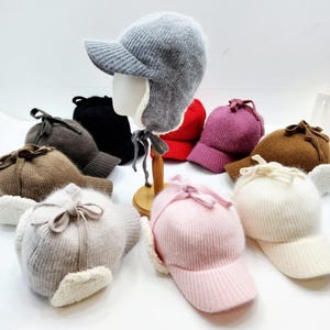 May include: A collection of winter hats in various colors, including gray, black, red, purple, brown, pink, and white. Each hat features a visor, ear flaps, and a decorative bow. The hats are made of a soft, textured material.