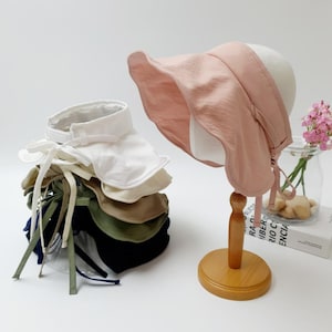 May include: A stack of six sun hats in various colors, including white, beige, green, and navy blue. The hats have a wide brim and a tie at the back. A pink sun hat is displayed on a wooden stand.