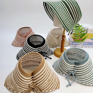 May include: A selection of sun visors in different colours, such as green and white stripes, brown and white stripes, black and white stripes, and tan and white stripes. Each visor has a wide brim and a tie closure.