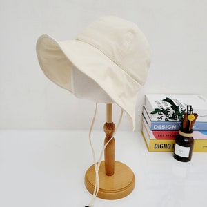 May include: A cream colored bucket hat with a wide brim and a long, cream colored drawstring. The hat is displayed on a wooden stand.