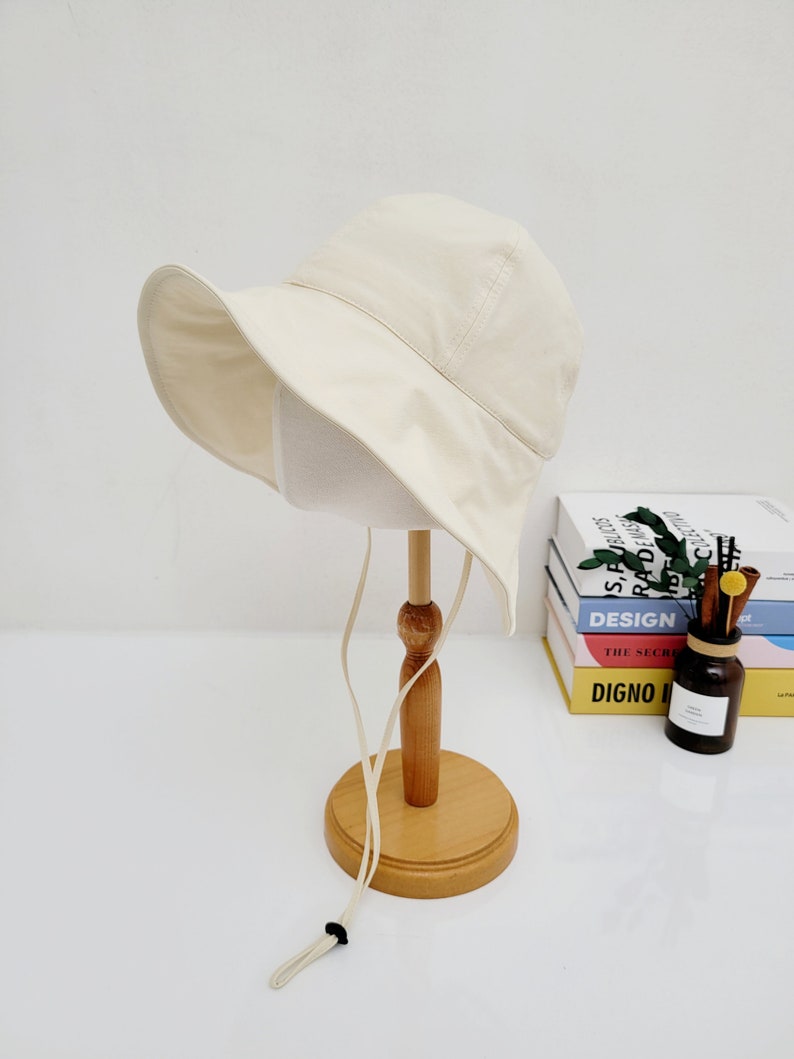 May include: A cream colored bucket hat with a wide brim and a long, cream colored drawstring. The hat is displayed on a wooden stand.