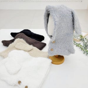 May include: A collection of fluffy, hooded hats with bunny ears in various colors: white, beige, brown, black, and gray. The gray hat is displayed on a mannequin head. Each hat features wooden button closures, offering a cozy and playful aesthetic.