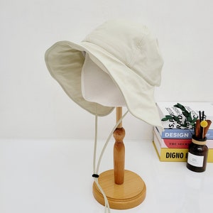 May include: A light beige bucket hat with a wide brim and a long, thin, light beige drawstring. The hat is displayed on a wooden stand.