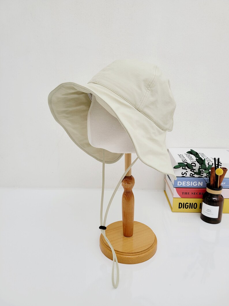 May include: A light beige bucket hat with a wide brim and a long, thin, light beige drawstring. The hat is displayed on a wooden stand.