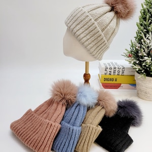 May include: A display of ribbed knit beanies in various colors, each topped with a fluffy pom-pom. The beanies include shades of pink, blue, tan, and black, with one displayed on a mannequin head. A small Christmas tree and books are in the background.