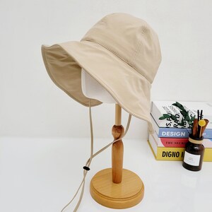 May include: A beige bucket hat with a wide brim and a long, adjustable strap. The hat is displayed on a wooden stand.