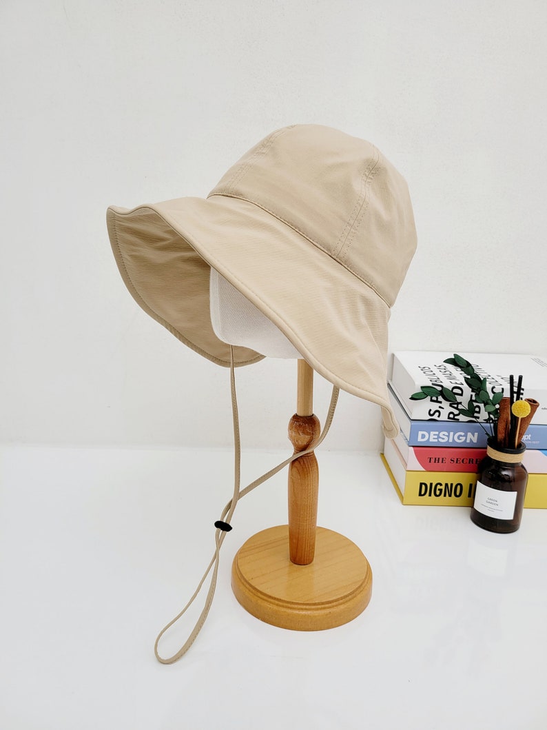 May include: A beige bucket hat with a wide brim and a long, adjustable strap. The hat is displayed on a wooden stand.