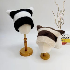 May include: Two striped furry hats with cat ears. The hat on the left is black and white, and the hat on the right is brown and white.