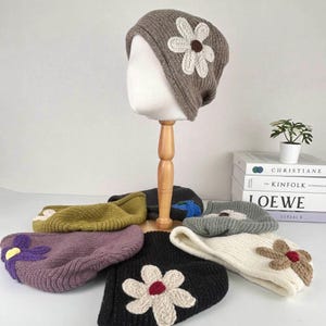 May include: A collection of knitted beanies, each adorned with a large, embroidered flower. The beanies come in various colors, including gray, black, purple, and olive green. One beanie is displayed on a mannequin head.