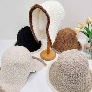 May include: A collection of fluffy bucket hats in various colors, including white, black, and shades of brown and beige. Each hat features a soft, textured material and a contrasting trim around the brim, offering a cozy and stylish accessory.