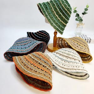 May include: A collection of handmade crochet bucket hats in various colors. The hats feature striped patterns in shades of green, blue, brown, and white. The hats are displayed on a white surface, showcasing their unique textures and designs.