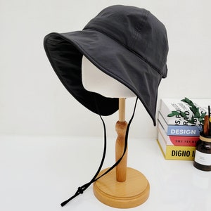May include: A black leather bucket hat with a wide brim and a chin strap. The hat is on a wooden stand.