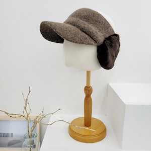 May include: A brown and white winter hat with ear flaps and a brim, displayed on a mannequin head. The hat is made of a textured fabric, likely wool or a wool blend. The ear flaps are a darker shade of brown.
