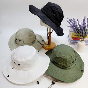 Puede incluir: Cuatro sombreros para exteriores en varios colores: negro, blanco, verde oliva y beige. Cada sombrero tiene un ala ancha, ojales y una correa ajustable para la barbilla. Los sombreros se muestran sobre una superficie blanca.