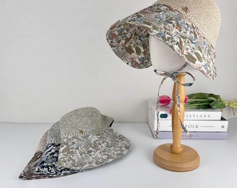 Puede incluir: Dos sombreros de sol de paja con bordes estampados florales. Un sombrero se exhibe en un soporte de madera, mostrando el diseño del borde de flores y hojas rosas, azules y verdes. El otro sombrero está plano, revelando un patrón floral similar y un patrón floral azul oscuro.