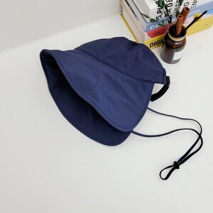 May include: A navy blue sun hat with a wide brim and a black adjustable strap. The hat has a fold-over design and a chin strap.