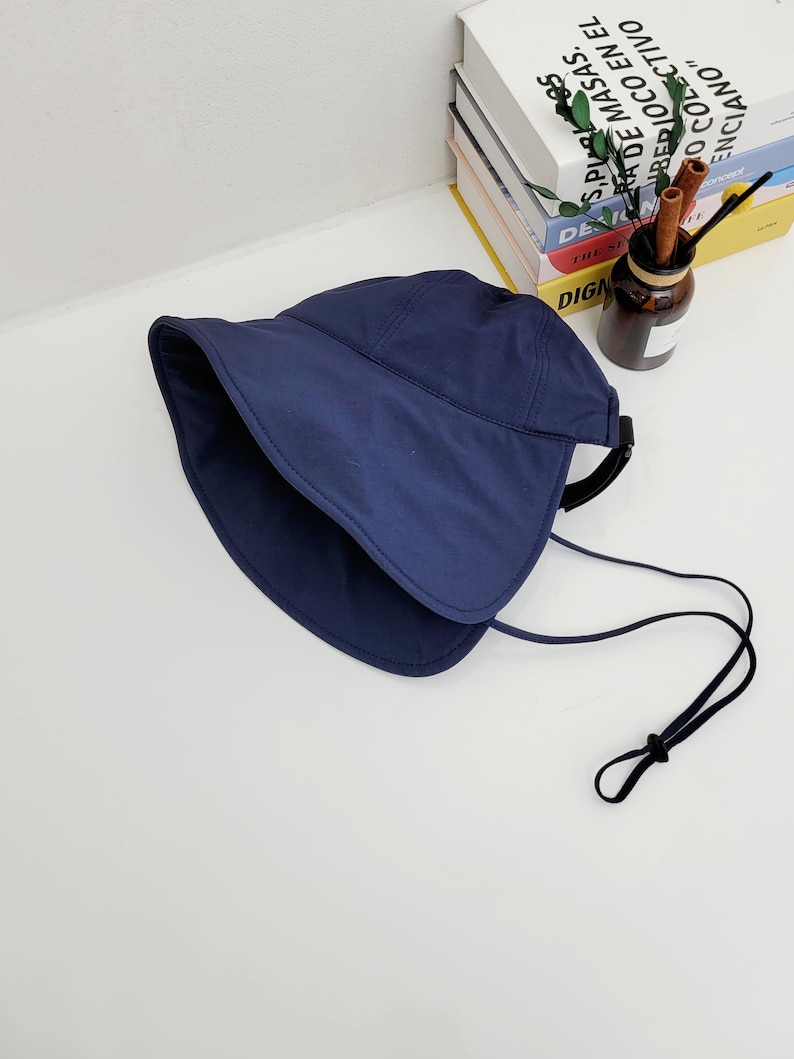 May include: A navy blue sun hat with a wide brim and a black adjustable strap. The hat has a fold-over design and a chin strap.