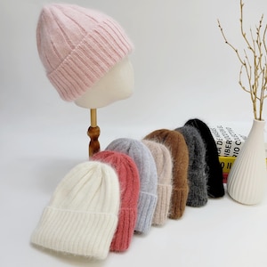 Peut inclure: Présentation de huit bonnets en tricot côtelé de différentes couleurs, dont rose, blanc, rouge et gris. Un bonnet rose est présenté sur une tête de mannequin. Les bonnets sont disposés en rangée, mettant en valeur leurs teintes et textures.