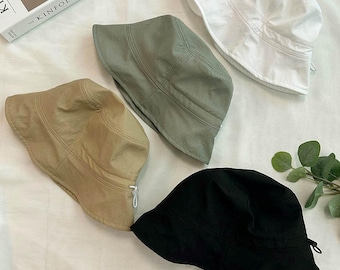 Könnte beinhalten: Vier Fischerhüte in verschiedenen Farben: Beige, Olivgrün, Weiß und Schwarz. Die Hüte sind aus leichtem Material gefertigt und haben ein schlichtes Design. Die Hüte sind auf einer weißen Oberfläche angeordnet.