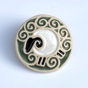 May include: A ceramic round ornament featuring a white and black stylised sheep design. The sheep is surrounded by a green swirl pattern.