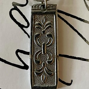 May include: A silver pendant necklace with a rectangular charm featuring an intricate floral design.
