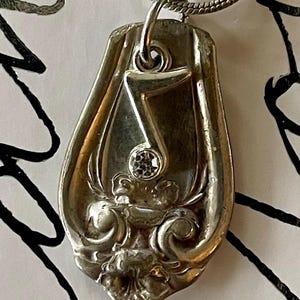May include: A silver spoon necklace with a number 7 charm and a small clear rhinestone. The spoon has a floral design.