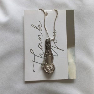 May include: A silver spoon necklace with a floral design and clear beads hanging from the spoon. The necklace is displayed on a white card with the text "Thank You" written in black cursive.