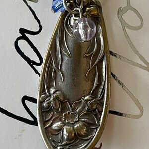 May include: A silver-toned pendant necklace featuring a spoon-shaped charm with floral detailing. The charm has a small blue and a lavender bead attached. The necklace chain is a silver color.