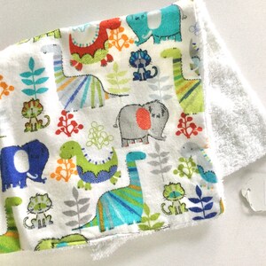 May include: White baby blanket with a colorful dinosaur, elephant, and cat print. The blanket is made of soft cotton and has a white terry cloth backing. The blanket has a tag that says "Muncie Market".