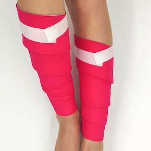 Può includere: Un paio di scaldagambe rosa con cinturini in velcro bianchi. Gli scaldagambe sono realizzati in un tessuto morbido ed elastico e sono progettati per essere indossati sopra leggings o pantaloni.