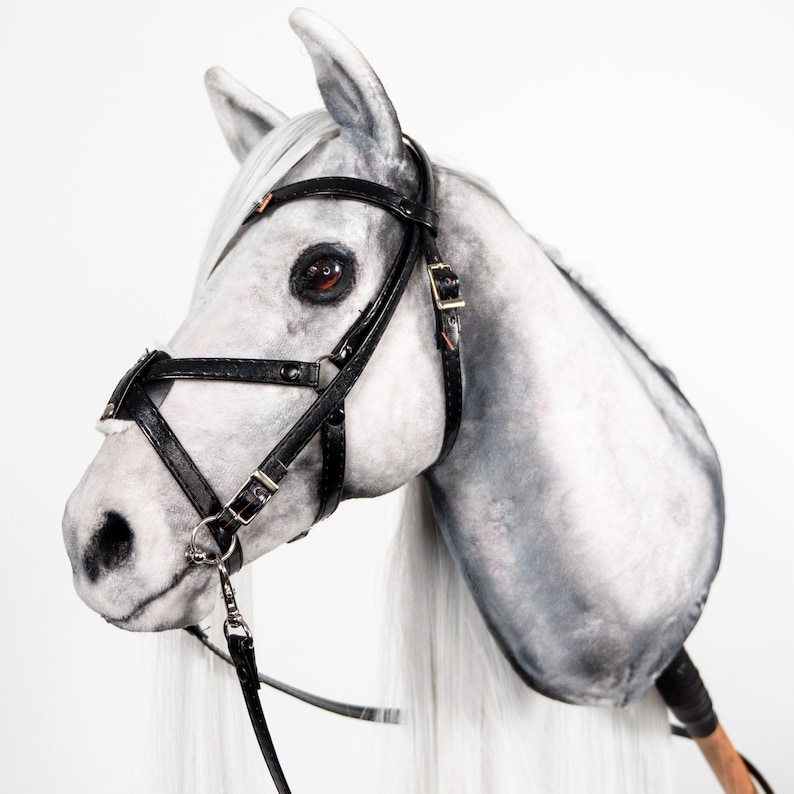 Pode incluir: Uma cabe&ccedil;a de cavalo de pel&uacute;cia branca com uma cabe&ccedil;ada e r&eacute;deas de couro preto. O cavalo tem uma longa crina e cauda brancas.
