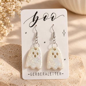 May include: A pair of white glittery ghost-shaped dangle earrings with silver-toned hooks. The earrings are displayed on a white card featuring the text "boo" in black script and "-GERBERALETTER-" at the bottom with small star icons.