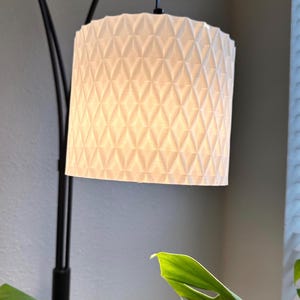 Puede incluir: Una pantalla de lámpara blanca con un patrón geométrico de diamantes cuelga de una lámpara de arco negra. La pantalla es cilíndrica y emite un brillo cálido. Una gran hoja verde de planta está en primer plano.