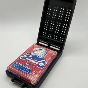 May include: Black plastic playing card case with a white clasp, holding a red, white, and blue Eagle Brand deck of cards. The case is open, revealing the cards and a perforated interior design.