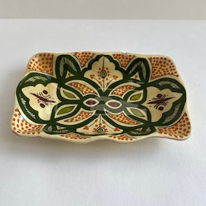 Op de afbeelding: Een rechthoekige keramische schaal met een geschulpte rand. De schaal heeft een crèmekleurige basis met een symmetrisch ontwerp in groen, oranje en bordeauxrood. Het ontwerp bevat bloemen en geometrische patronen en oranje stippen.