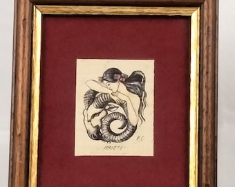 May include: A framed artwork featuring a pen and ink drawing of a woman with long hair embracing a ram. The artwork is set against a burgundy background within a wooden frame with gold trim. The word "ARIETE" is written below the drawing.
