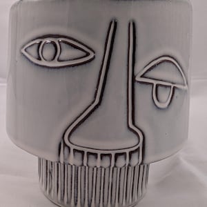 May include: A ceramic planter with a face design. The light gray planter features a stylized face with dark brown outlines. The eyes, nose, and mouth are depicted with simple lines. The base has vertical ridges.