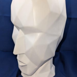 May include: A white, geometric-shaped head sculpture with a detailed, angular design. The sculpture features a prominent nose, defined lips, and a smooth, matte finish. The head is set against a dark blue background.