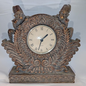 May include: A decorative quartz clock with a bronze finish. Two monkey figures flank the clock face, which features Roman numerals. The clock has an ornate, carved design and sits on a rectangular base.