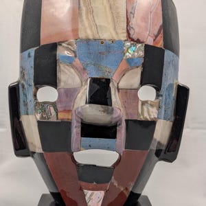 May include: A decorative mask sculpture crafted from inlaid stone and shell segments in black, blue, tan, and reddish-brown tones. The face features geometric patterns, cut-out eyes and mouth, and a small blue sticker reading Hecho en Mexico.