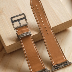 May include: Two tan leather watch bands with white stitching and silver-tone hardware. The bands are displayed on a light wooden surface. The bands are designed for a smartwatch.