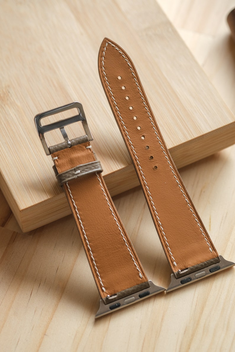 May include: Two tan leather watch bands with white stitching and silver-tone hardware. The bands are displayed on a light wooden surface. The bands are designed for a smartwatch.