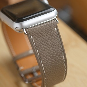 May include: A silver Apple Watch with a taupe leather band. The band has white stitching and a silver buckle. The watch face is black. The leather has a textured appearance.
